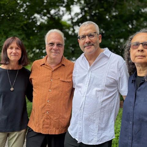 Group portrait of four people standing side by side on a green lawn, casually dressed, with trees in the background.