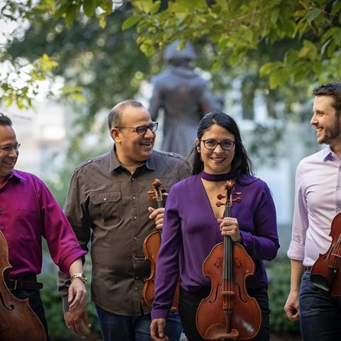 Four adults standing outdoors, posing together in front of leafy trees, smiling or looking at the camera each holding their instrument.