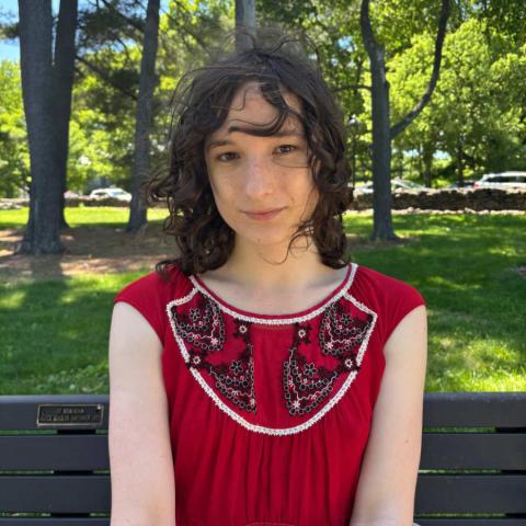 A photo of Mae Buck ’26, a person with long brown hair and a red dress. Buck is sitting outside in the sunlight with grass in the background.