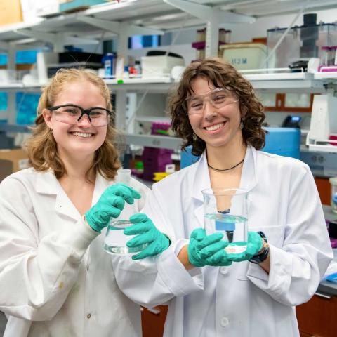 Two smiling people in a lab wearing gloves and holding lab containers.