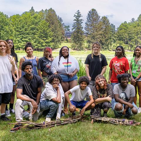 A diverse group of fifteen people is posing for a photo on a grassy lawn with a pond and a college building visible in the background.