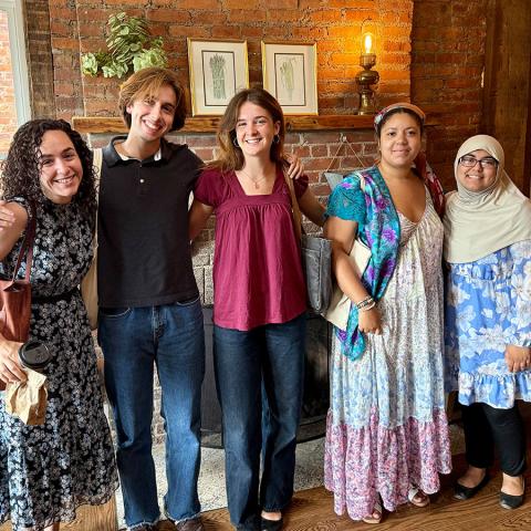 A professor and four students, all smiling, pose in front of a brick fireplace.