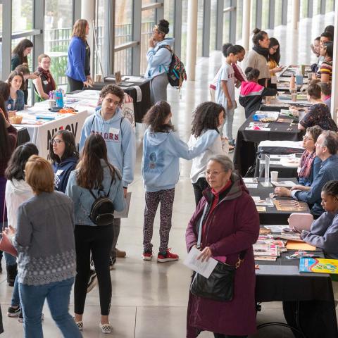 People gather at an indoor event, walking among rows of tables where representatives share information and materials.
