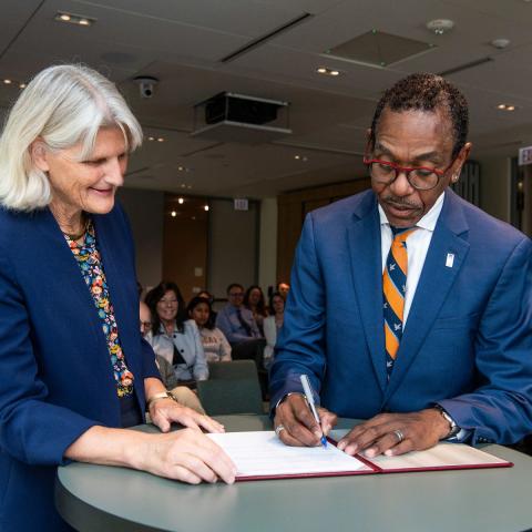 Two people signing a document at a table, with others seated in the background.