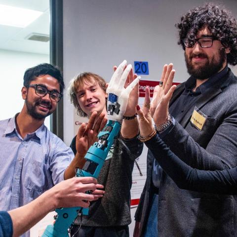 A group of onlookers checking out a robotic hand, held by a person, offerring a high five.
