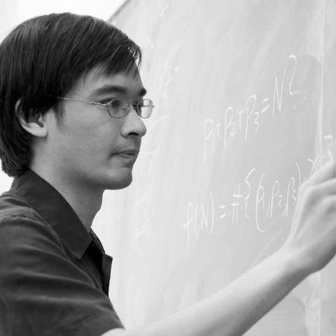  Black-and-white photo of a person writing mathematical formulas on a chalkboard.