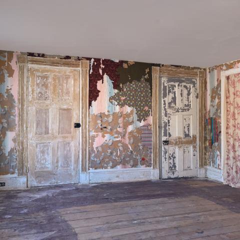 Installation view showing layered mixed textiles integrated with existing wallpaper in a weathered interior room.