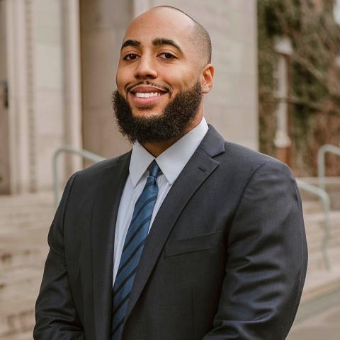 Philip McHarris with a neatly groomed beard wearing a dark suit, light blue shirt, and striped tie, smiling while standing outdoors in front of stone steps and a building.