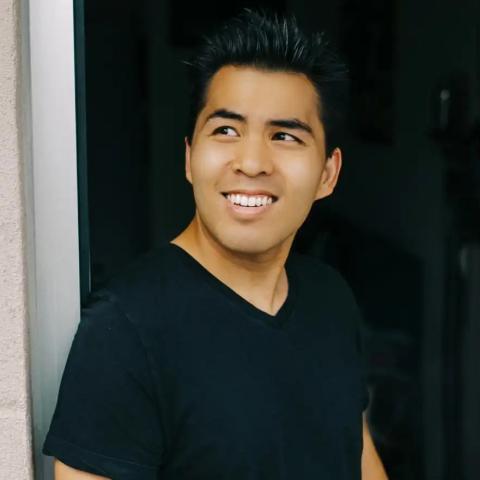 A person with short dark hair wearing a black T-shirt stands in a doorway, smiling and looking to the side.