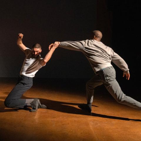 Two dancers perform on stage; one kneels with an arm raised while being pulled forward by the other, who leans away with an outstretched arm.