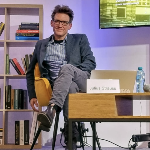 Julius Strauss, a person with short, tousled dark hair and glasses, is seated in a mustard-yellow chair with one leg crossed over the other. They wear a dark blue blazer, a striped shirt, grey pants, and black boots. To their right is a wooden table with a name card that reads "Julius Strauss," a plastic water bottle, and a microphone. Behind them is a bookcase filled with colorful books, and a screen displaying their photo, name, and the title "Ex war correspondent."