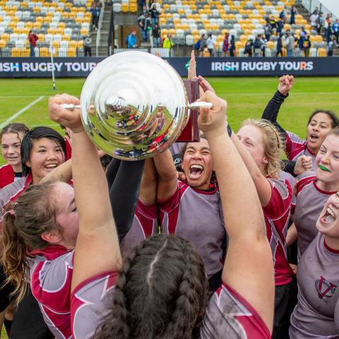 Vassar College Women’s rugby team in maroon and gray jerseys celebrating on the field, holding up a large silver trophy together with excitement.