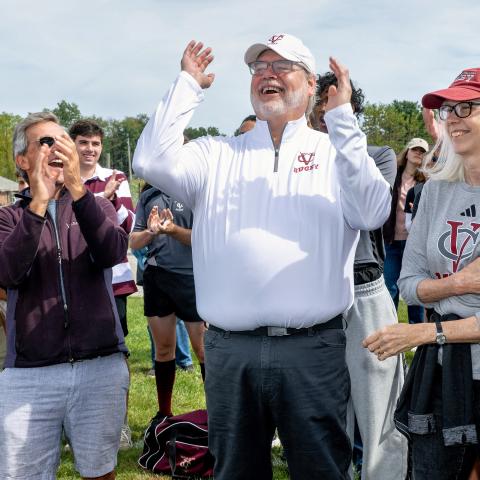 A crowd stands outdoors on grass, clapping and smiling. In the center, a person in a white quarter-zip pullover with a maroon “Rugby” logo raises both arms joyfully. People around them, some wearing sunglasses and Vassar-themed clothing, join in the celebration. The background includes trees and people partially visible in athletic uniforms.