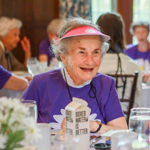 June Ross Marks ’49 with short white hair wearing a bright purple shirt and a pink visor, seated at a dining table.