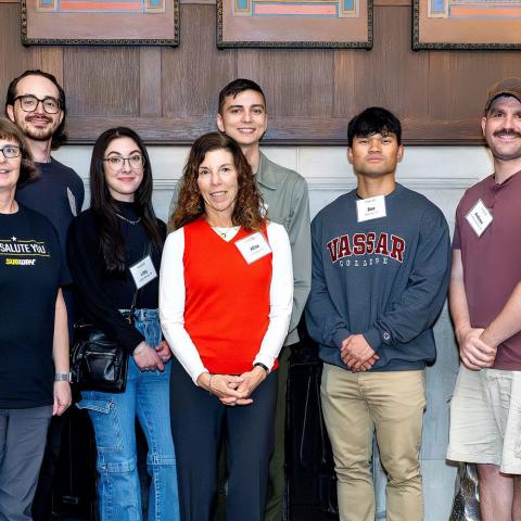 Eleven people stand in a line indoors, smiling at the camera, wearing name badges; one in a “We Salute You” T-shirt, one in a Vassar sweatshirt, one in a red sweater, with wood-paneled walls and framed art behind them.
