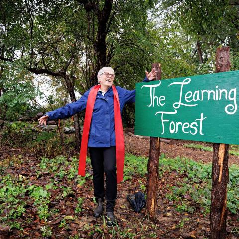 A smiling older woman with short gray hair, wearing a bright blue jacket and a long red scarf, stands with her arms outstretched in a lush, wooded area. To her right is a green wooden sign on two rustic posts that reads "The Learning Forest" in white script.
