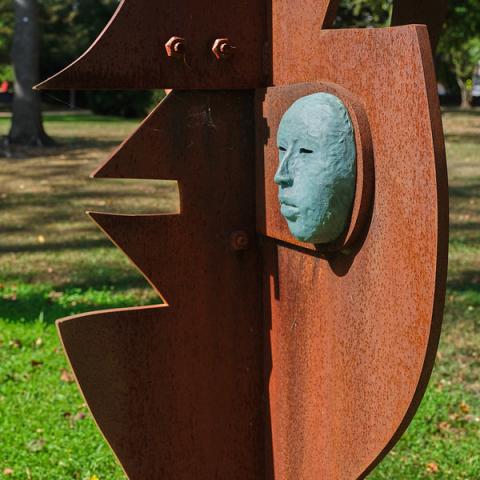  A close-up of a rustic metal sculpture with a weathered, rusted surface. A small, green-patina mask of a human face is embedded in a cutout of the rusty metal. The sculpture is outdoors, with green grass and trees visible in the background.