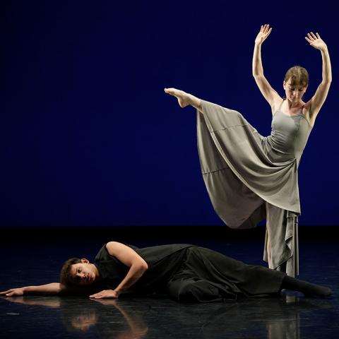 Two contemporary dancers on a dim stage: one in black lies on the floor, arm extended; behind, a dancer in a flowing gray dress stands with arms raised and one leg lifted in a side extension against a deep blue backdrop, their reflections visible on the glossy floor.