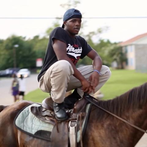 A a still from the film Hale County This Morning, This Evening featuring one of the subjects, a person crouching while standing on the saddle of a horse.