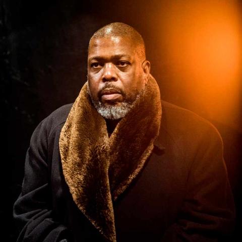 A close-up, dramatic portrait of a person with a gray beard and short hair, looking intently into the distance. The person is wearing a dark coat with a wide, brown faux-fur collar, black gloves, and a striped cuff visible. The background is dark, with a warm orange glow in the top right.