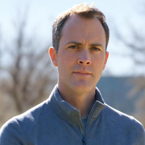 Outdoor head-and-shoulders portrait of a person in a gray quarter-zip, neutral expression, with blurred trees and a blue building behind the person.