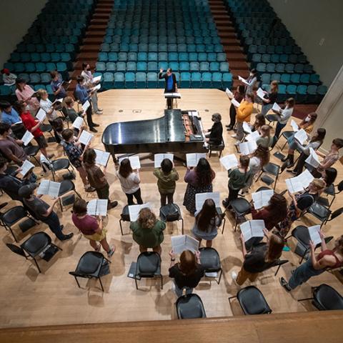 Overhead view of a choir holding sheet music and forming a semicircle around a grand piano on stage. A conductor stands at the front, with rows of empty teal seats filling the auditorium behind.
