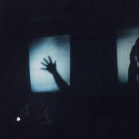A darkened scene features two illuminated rectangular screens side by side against a black background. The left screen shows the shadow of an outstretched hand and forearm, angled upward with fingers splayed. The right screen displays the shadowy silhouette of a figure bent forward, hands clasped low and head down, partially obscured by darkness. Two small desk lamps sit below the screens, casting faint light upward.