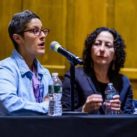 A person with short hair and glasses speaks into a microphone at a panel, wearing a light blue shirt. Another person is seated next to them.