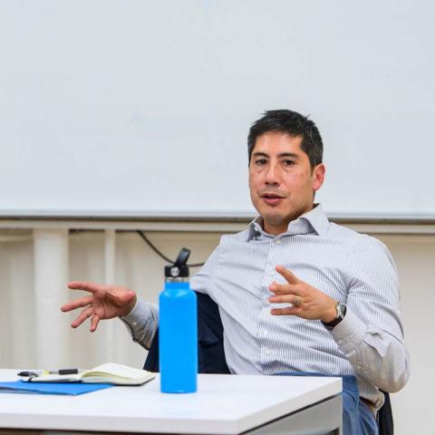 A person seated at a white table, wearing a light blue striped dress shirt and gesturing with both hands while speaking. A bright blue water bottle, a small notepad, and a pen are on the table. A blank white board is visible in the background.