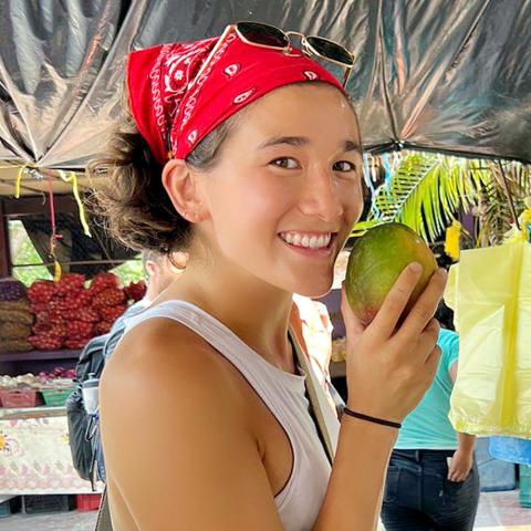 Smiling shopper in a red bandana and sunglasses holds a mango at an outdoor market, standing under a tarp beside hanging yellow and blue bags, with produce stalls and palm fronds in the background.