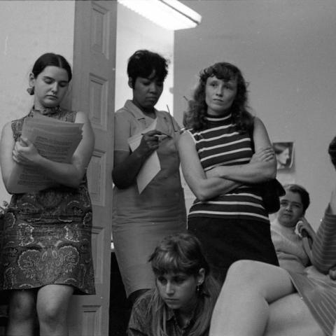 Several individuals gather closely in a small room with plain white walls and a fluorescent ceiling light. Some are seated while others stand, holding or reviewing stacks of typed pages, with one person in the center crossing their arms and another writing on a pad near the doorway. Attentive expressions are visible throughout the group, with a mix of seated and leaning postures. A tall shelf filled with papers stands to the left, and a small framed portrait hangs on the back wall.