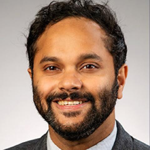 Close-up professional headshot of an individual with dark hair and a full beard, smiling slightly. They are wearing a dark suit jacket over a light-colored collared shirt, against a plain gray background.