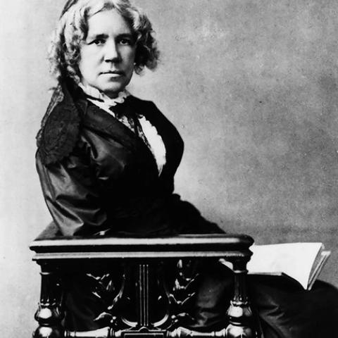 Black-and-white studio portrait of a seated woman in a dark dress, curled hair, papers at her side on a carved chair.
