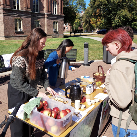 A table is set up outdoors, featuring various food items, including a plastic container filled with red apples and baked goods. Two individuals are engaged in preparation; one is pouring from a metal kettle, while the other places items on the table. In the foreground, a person with short, colorful hair is observing the table and holding a cup. The setting is vibrant, characterized by sunlight casting soft shadows, and the surrounding area displays green grass and trees with autumn foliage.