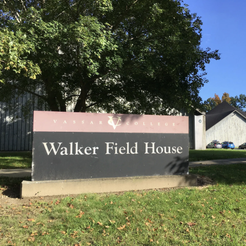 A large sign stands prominently on a grassy area, displaying the words "Vassar College" in bold lettering at the top, followed by "Walker Field House" in smaller text below. The sign has a dark background with a maroon strip at the top, contrasting with the bright green grass and scattered fallen leaves in the foreground. Behind the sign, a partially visible, multi-story building features a light-colored wooden facade, with trees and a clear blue sky above creating a vibrant atmosphere.