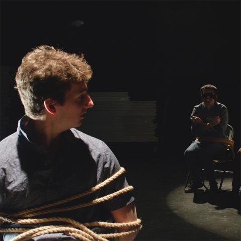 In a dark stage setting, a young man in the foreground is tied with thick rope around his torso and arms, looking left, while another person sits bound to a chair in a spotlight in the background.