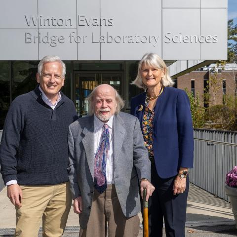 Three adults stand smiling in front of a campus building sign that reads “Winton Evans Bridge for Laboratory Sciences”; the man in the center holds a cane, pink mums in planters flank the walkway, and fall trees are visible in the background.