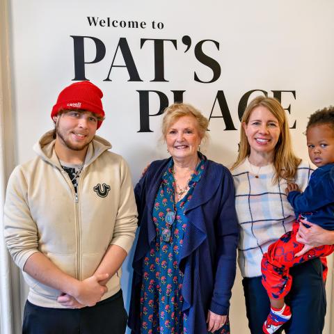 Three adults and one child pose indoors in front of a wall sign reading ‘Welcome to Pat’s Place’.