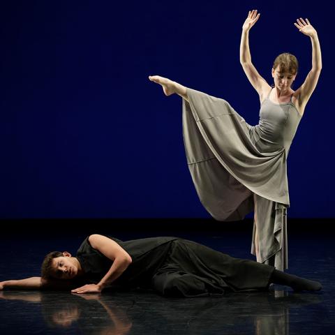 Two dancers on a stage with a dark blue backdrop: one lies on the floor in black while the other stands in a gray dress with one leg lifted high and both arms raised.