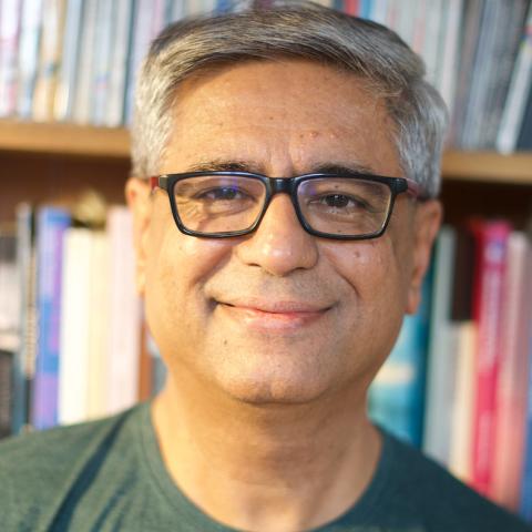 Smiling person with short gray hair and black glasses, wearing a green T-shirt, in front of a bookshelf with colorful books.