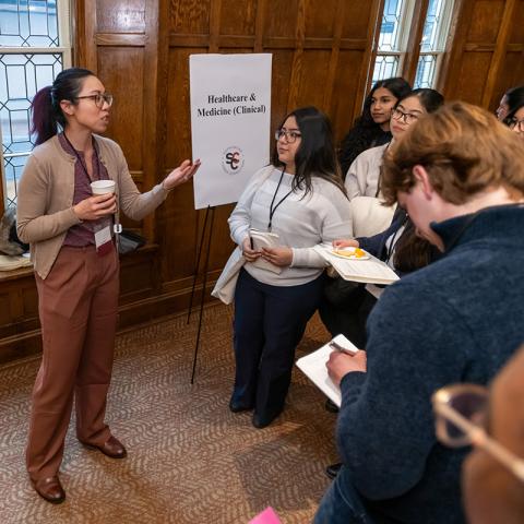 A speaker gestures while addressing a small group of students wearing name badges at a networking event near a “Healthcare & Medicine (Clinical)” sign.