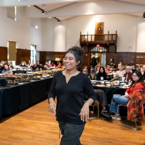 A smiling attendee walks through a large hall as seated guests watch, with tables of food and a wood-paneled room in the background.