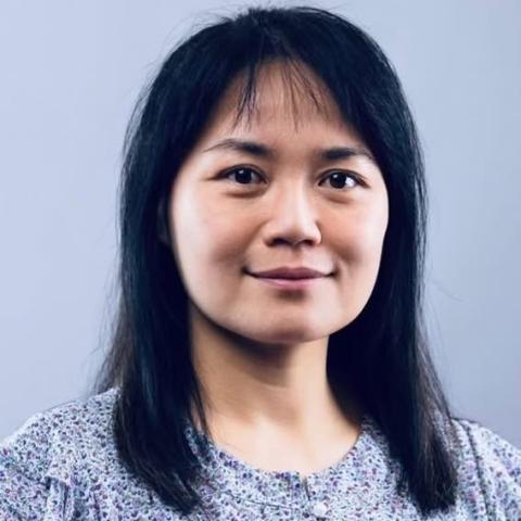 Headshot of a person with straight dark hair and bangs, wearing a patterned blouse, looking at the camera with a slight smile against a plain light gray-blue background.