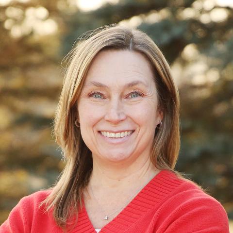 Amy Allen stands outdoors with her arms crossed, smiling, wearing a red sweater against a softly blurred natural background.