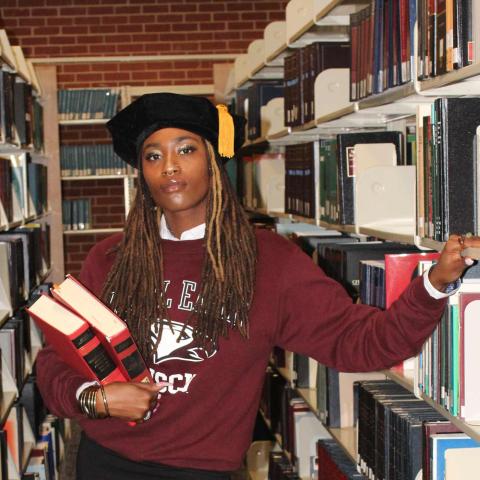 An individual wearing a maroon sweatshirt, black skirt, and academic cap with a yellow tassel stands in a library aisle. They hold two red books under one arm while resting their other hand on a bookshelf.