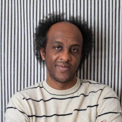 A person with dark curly hair and a slight smile poses with arms crossed. They wear a light-colored sweater with thin horizontal black stripes against a background of vertical black and white stripes.