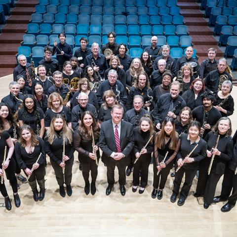 A group portrait of the wind ensemble members with instruments in concert dress on the stage.