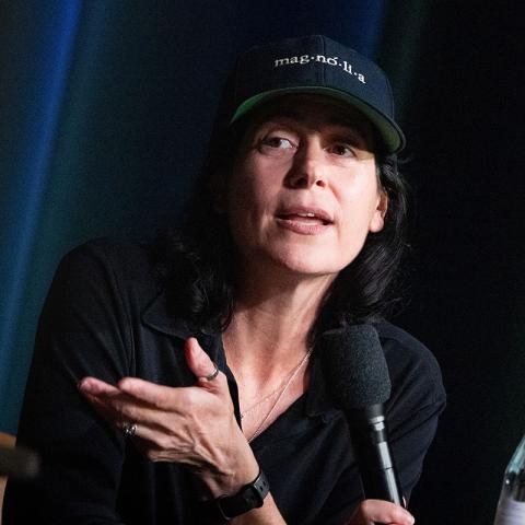 A person wearing a dark shirt and a "mag·no·li·a" baseball cap speaks into a microphone while gesturing with their right hand.