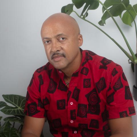 A portrait of an individual wearing a red and black patterned shirt, sitting between two potted plants against a neutral white wall.
