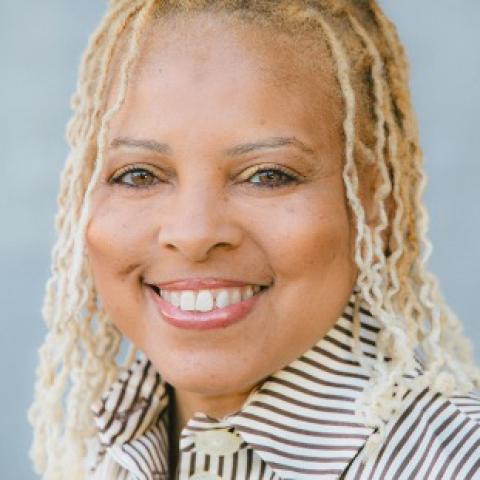 Smiling person with light blond locs wearing a striped collared shirt, against a soft blue background.